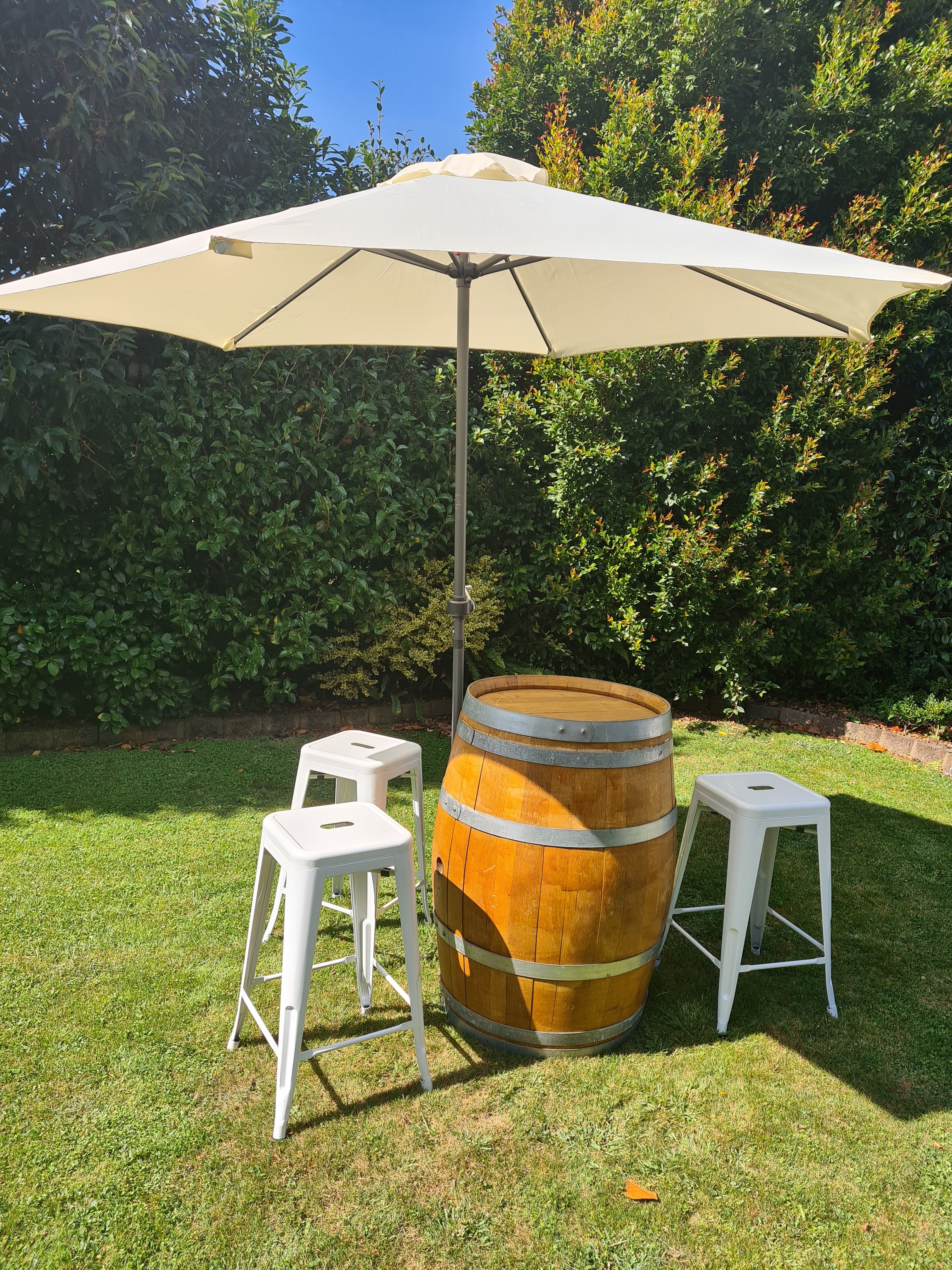 Wooden barrel with white stools and a beige umbrella on grass